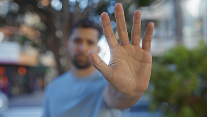 Man standing outdoors in urban park environment with blurred background extending hand to camera in gesture emphasizing focus on palm against city street.