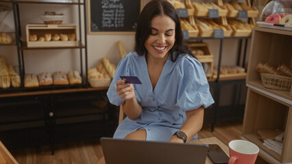 Woman shopping online with a credit card in a bakery, wearing a blue dress, smiling, sitting surrounded by bread and pastries, using a laptop, interior setting, daytime.