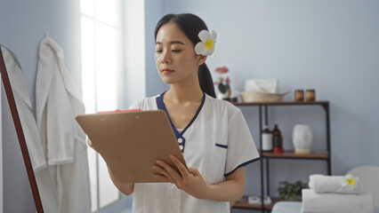 Asian woman in a spa room with wellness equipment checks a clipboard, wearing a flower in her hair,...