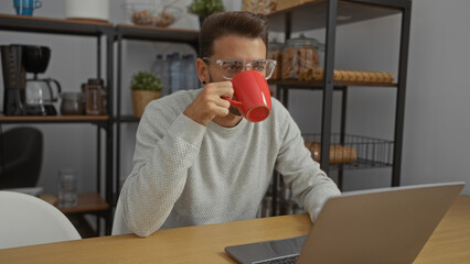 Young man in office sipping coffee from a red mug while working on a laptop at a modern workplace with shelves in the background, creating a casual and productive atmosphere.