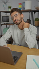 Young hispanic man seated at an office desk working on a laptop with glasses feeling neck pain...