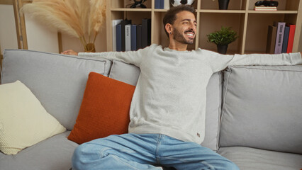 Obraz premium Young man relaxing on a comfortable sofa in a modern home living room, surrounded by books and plants, showcasing tranquility and casual lifestyle indoors.