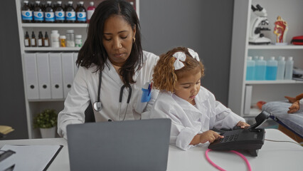 Woman doctor with girl in clinic room working on laptop and phone indicating a busy pediatric...