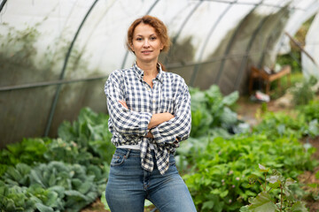 Girl worker shirt is standing in greenhouse with rake and other tools in hands. She is resting...