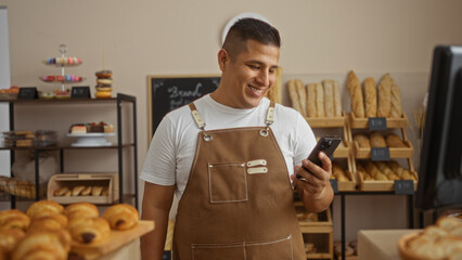Young hispanic man in a bakery shop looking at his phone while smiling, wearing a brown apron,...