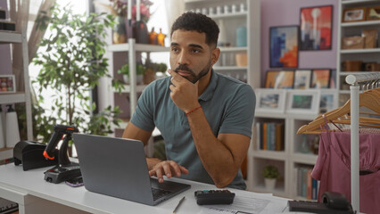 Young man thinking while working on a laptop in a home decor store with various items like clothes...
