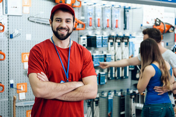 Confident smiling salesman on foreground in power tools store. Guy is ready to help clients on background.