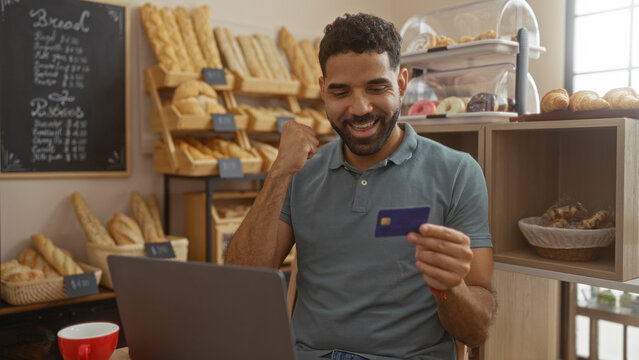 Young man in a bakery excitedly looking at a credit card while using a laptop with bread and pastry shelves in the background.