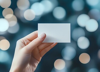 Female hand holding blank white business card against blurred bokeh lights background in blue tones, mockup for branding and corporate identity design.