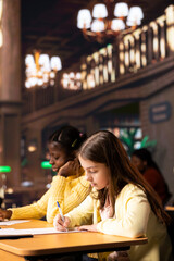 Diverse schoolgirls taking notes and completing theoretical exercises in a public library, showcasing positive academic development and a productive homework session. Scholars solve assignments.