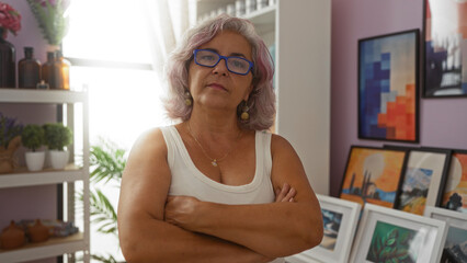 Middle-aged hispanic woman with gray hair and glasses standing confidently with arms crossed in front of a pink wall background with colorful framed art behind her.