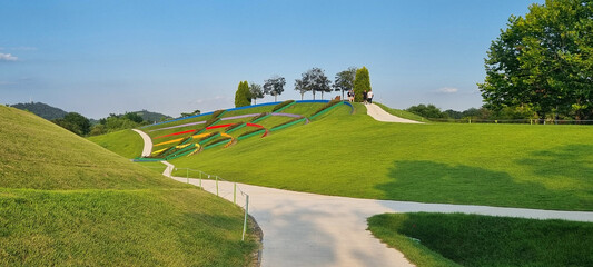 Side view of a colorful striped hill at International Garden Expo, Suncheon, South Korea, with sandy walking paths, green grass, and people strolling under a blue sky.