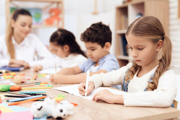 Children write in notebooks with a pen. They are sitting at the table. They are helped by an adult teacher.