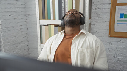 Young man with headphones relaxes in an office, leaning back in a chair, eyes closed, enjoying music, surrounded by books and a casual atmosphere.