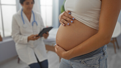 Pregnant woman with belly in a clinic room while doctor sits with a tablet providing medical...