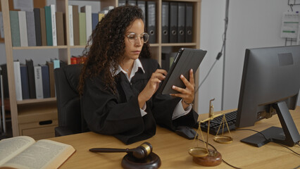 Woman judge with curly hair using tablet in office surrounded by books and judicial tools, focusing...