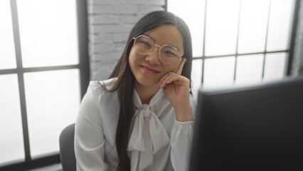Fototapeta premium Young beautiful asian woman in office setting with glasses sitting in modern interior workplace smiling confidently at camera.
