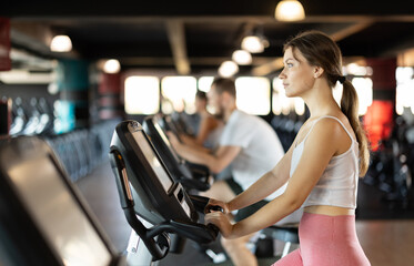 Fototapeta premium Girl performs exercises with stationary bicycle. Female gym visitor performs series of repetitions movements on simulator. Shot of sexy muscular woman in sportswear and good physique