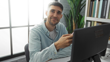 Fototapeta premium Young hispanic man working in modern office using computer, wearing casual shirt, smiling confidently with bookshelf and large window in background, bright and inviting ambiance