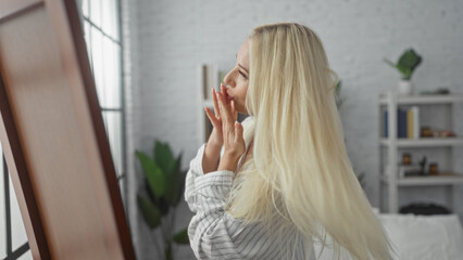 Fototapeta premium Woman with blonde hair enjoying morning indoors in a modern bedroom, reflecting in a mirror while wearing a striped shirt, exuding elegance and serenity within a cozy home setting.