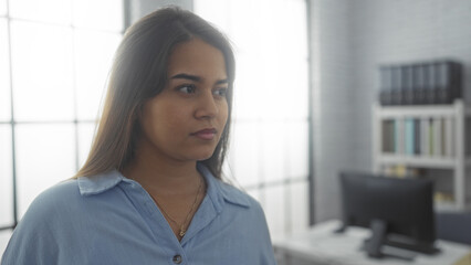 Young woman in a modern office setting, looking thoughtful and focused with natural light filtering through large windows, emphasizing a professional and calm atmosphere.