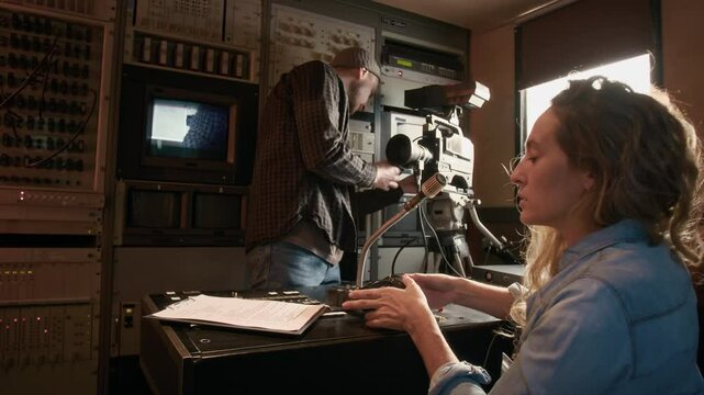 Medium full shot of young Caucasian female journalist putting on press pass and reading script, while preparing to present news piece, and male operator setting up video camera, in TV production truck