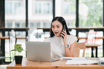 Fototapeta premium A young woman enthusiastically talks on her smartphone while working on a laptop in a bright, modern office setting.