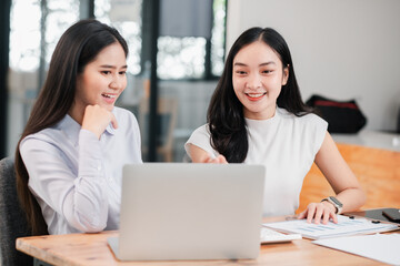 Two young women working together in a modern office, discussing ideas and using a laptop for their project.