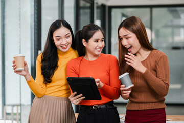 Three young women in a modern office setting, sharing coffee and using a tablet, smiling and interacting.
