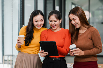 Three women in a modern office setting, smiling and using a tablet while holding coffee cups, showcasing a casual and collaborative work environment.