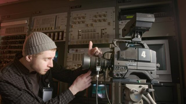 Medium shot of young Caucasian male operator in checked shirt and beanie setting up video camera in TV truck, adjusting tripod position, checking lens, while preparing to broadcast news report