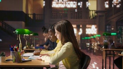 Preteen girls focus on reading the required books at a study desk, developing their academic skills and literary language at the library. Scholars enjoy lecture for the school curriculum. Camera A.