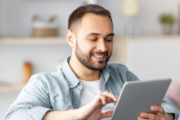 Cheerful young man using tablet pc, working or studying online from home. Portrait of positive Caucasian guy making business project, having video call or remote meeting indoors