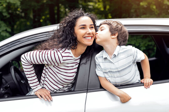 Cheerful little arabic boy kisses his mother on cheek through open window in new car, enjoy journey together, outdoor. Summer trip with family, love and family relationship, auto tour at vacation