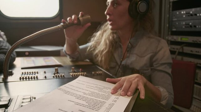 Medium closeup of Caucasian female journalist with headphones sitting in TV van, tapping on microphone, checking sound, talking to remote production crew before going live with report