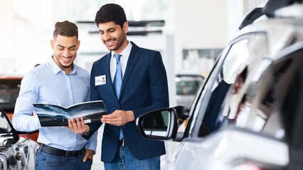 Middle-eastern guy having conversation with sales assistant in auto showroom. Handsome car dealer...