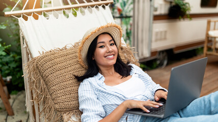 Joyful Asian lady using laptop computer while lying in hammock near RV on autumn camping trip. Cheery young woman working or studying online, browsing web outdoors on weekend