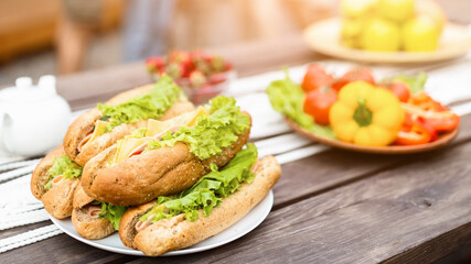 Sandwiches and vegetables, picnic foods on wooden table outdoors, group of young friends toasting with beer on background, selective focus. Copy space. Autumn camping trip concept