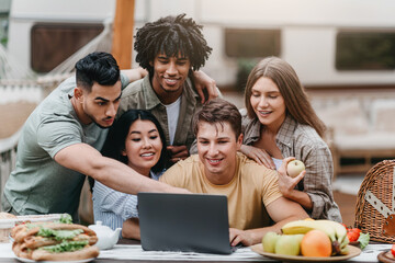Group of interracial young people using laptop while camping in countryside, video chatting online, having fun. Diverse millennial friends spending time together outdoors during summer holidays