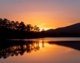 Obraz premium A stunning sunset over a calm lake with silhouettes of trees and mountains reflecting on the water.