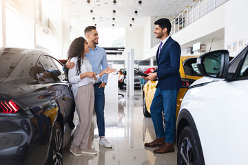 Wealthy middle eastern young couple having conversation with good-looking sales assistant at luxury auto showroom, looking for new car, gesturing and smiling, full length photo, panorama
