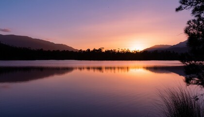 Fototapeta premium A stunning sunset over a calm lake with silhouettes of trees and mountains reflecting on the water.
