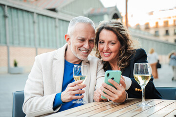 Joyful Mature Couple Sharing Special Moments While Engaging With Their Smartphone And Enjoying Delicious White Wine Together At A Cozy Table In An Outdoor Setting Filled With Warm Smiles And Laughter