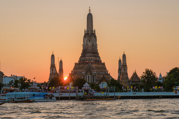 Fototapeta premium Golden rays of the setting sun illuminate the majestic Wat Arun temple, casting stunning reflections on the Chao Phraya River in Bangkok. The vibrant hues create a magical atmosphere.