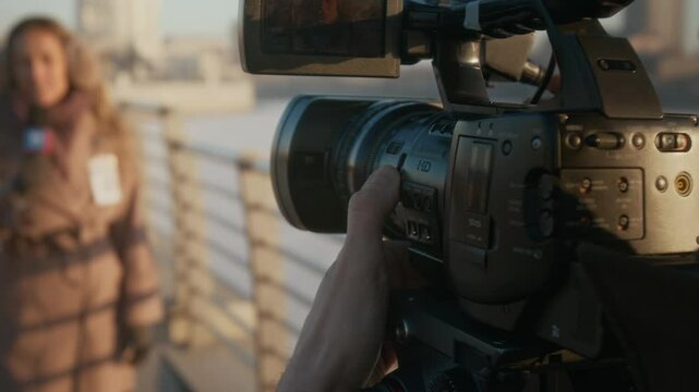 Over-shoulder shot of hands of unrecognizable male TV operator opening camera lens, setting up focus then signaling female reporter to begin speaking, while recording news item on city river bridge