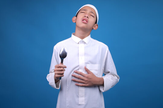 Unhappy Young Muslim Boy Touching His Stomach Feels Hungry Isolated on Blue Background. Ramadan and Eid Fitr Concept.