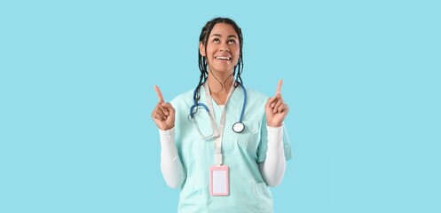 Young African-American female doctor pointing upwards on light blue background with space for text