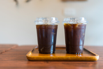 Iced Americano filled with ice and black coffee in a transparent take out plastic coffee cup on a wood tray, two glass