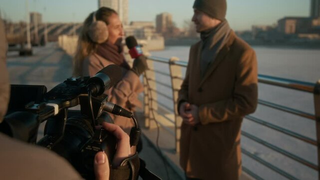 Over-shoulder shot of anonymous TV operator with camera filming young Caucasian female reporter talking to male local council head in smart winter coat while standing on river bridge