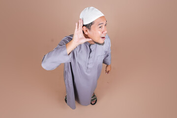High Angle of Young Muslim Man Heard Something Interesting Holding Hand to Ear Isolated on Beige Background